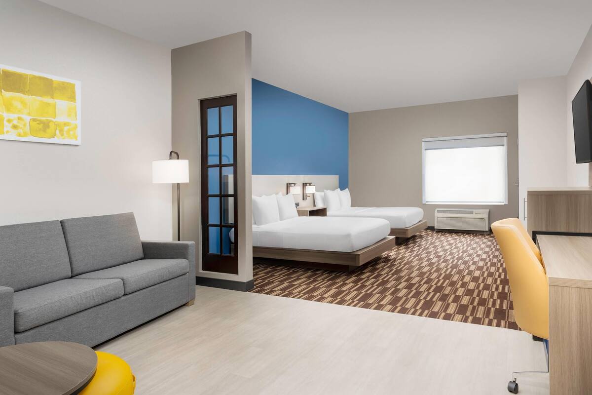 A hotel room with a gray sofa, a bed with white linens, a desk with a chair, and a window with blinds.