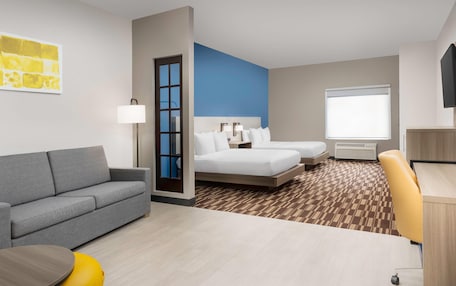 A hotel room with a gray sofa, a bed with white linens, a desk with a chair, and a window with blinds.