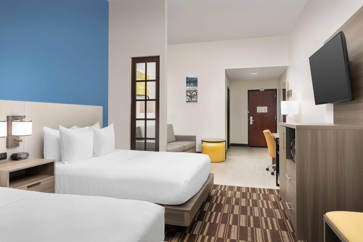 A hotel room with two beds, a desk, a chair, a TV, and a small table with a lamp.