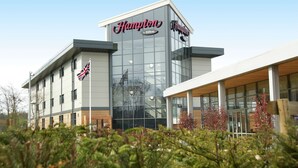 A Hampton by Hilton hotel with a modern glass facade, a flag, and a sign indicating it's part of the Hilton brand. - Hampton by Hilton Corby/Kettering (Corby)