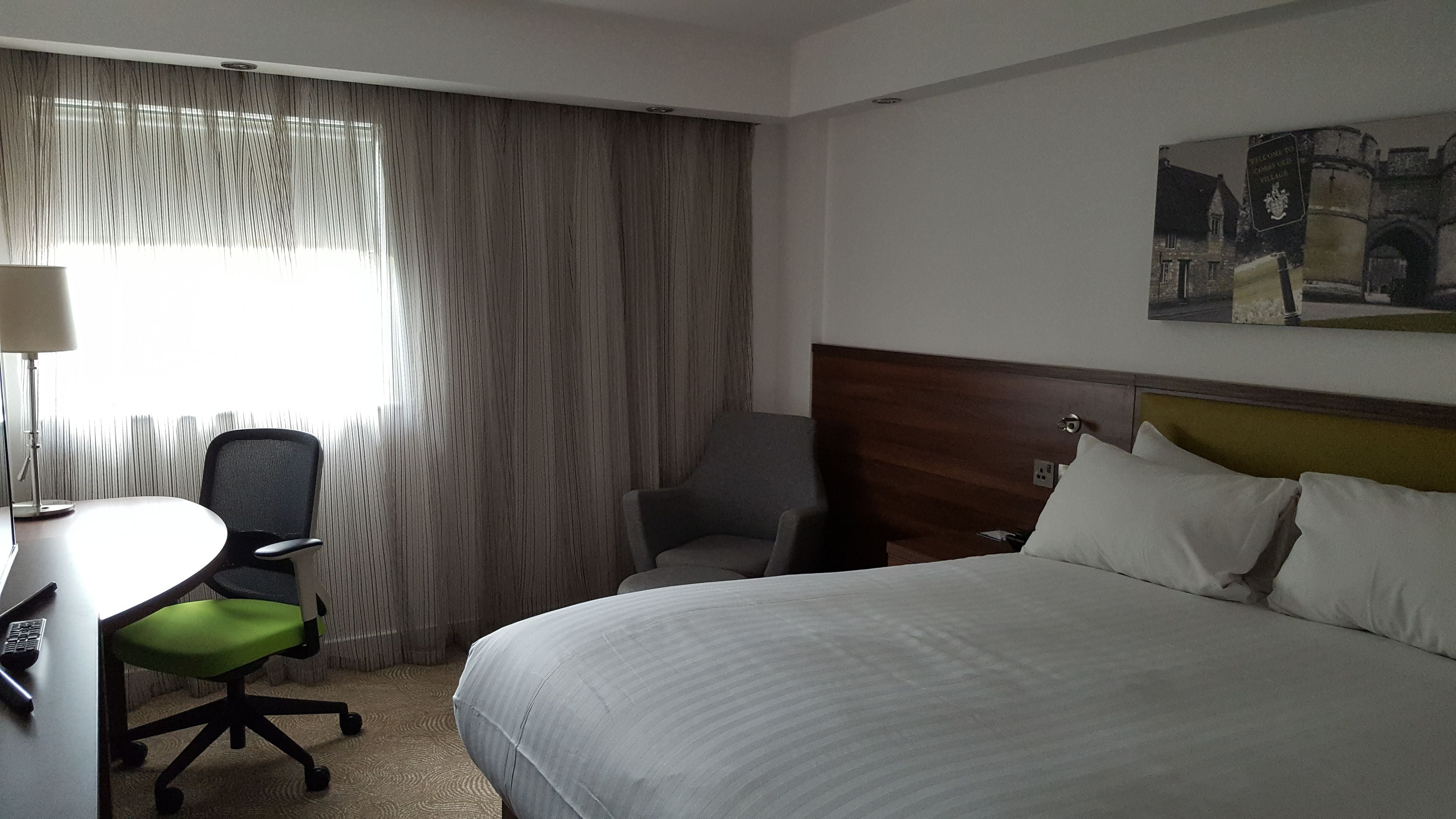 A hotel room with a bed, a desk with a chair, a lamp, and a painting on the wall.