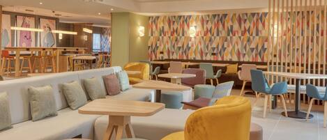 A modern interior with a seating area featuring sofas and round tables, a colorful geometric wall, and pendant lighting.