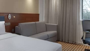 A hotel room with a bed, a grey sofa, a desk with a chair, a window with curtains, and a patterned carpet.
