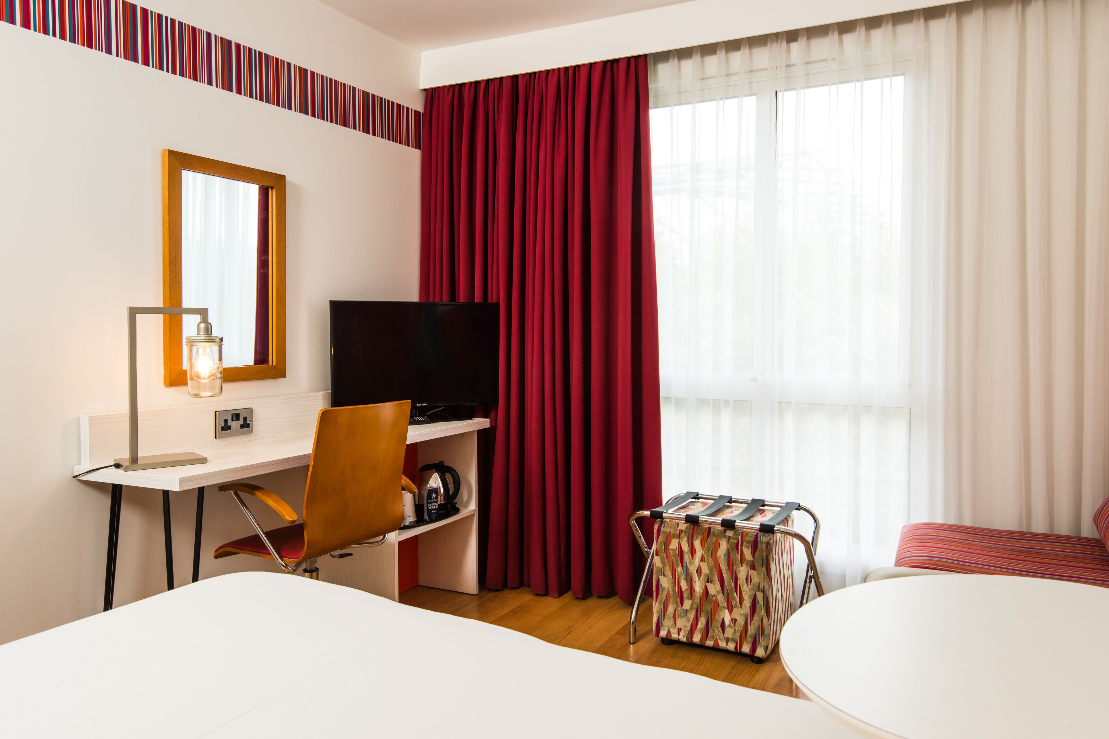 Ein Hotelzimmer mit Schreibtisch, Stuhl, Fernseher und einem kleinen Tisch mit einem Koffer.