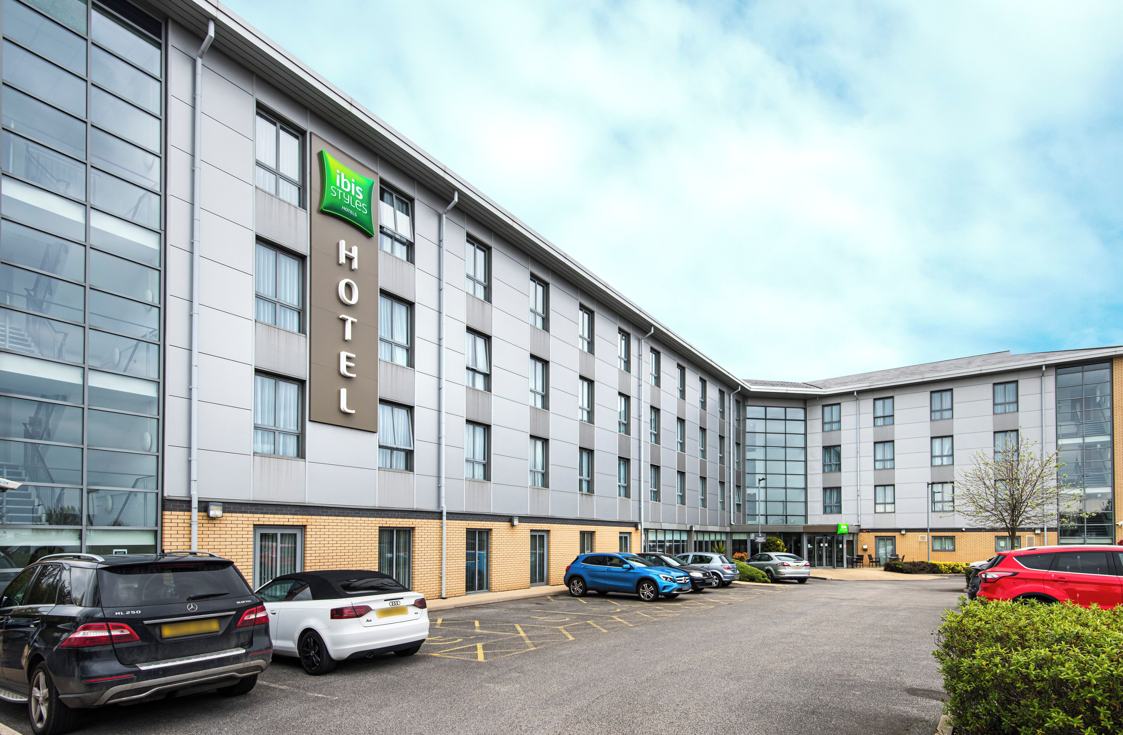 A modern hotel building with a sign for 'Ibis Styles Hotel'. There are several cars parked in front, and the building has a large glass facade.