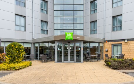 A modern hotel entrance with a glass facade, outdoor seating area, and a sign for ibis Styles hotel.