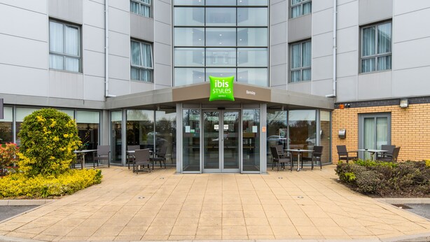 Een moderne hotelentree met een glazen gevel, een terras en een bord met de tekst ibis Styles.