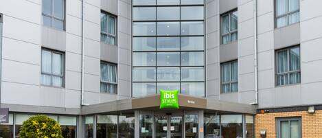 A modern hotel entrance with a glass facade, outdoor seating area, and a green sign indicating the hotel name.