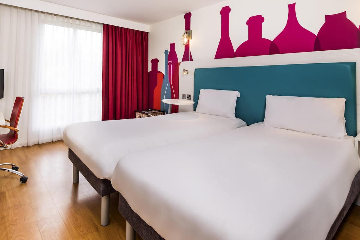 Une chambre d’hôtel avec deux lits, un bureau, une chaise et un mur décoré d’œuvres d’art abstraites.