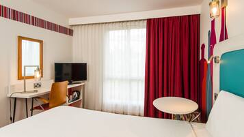 Habitación de hotel con cama, escritorio con televisor, una silla, una mesita y una ventana con cortinas.