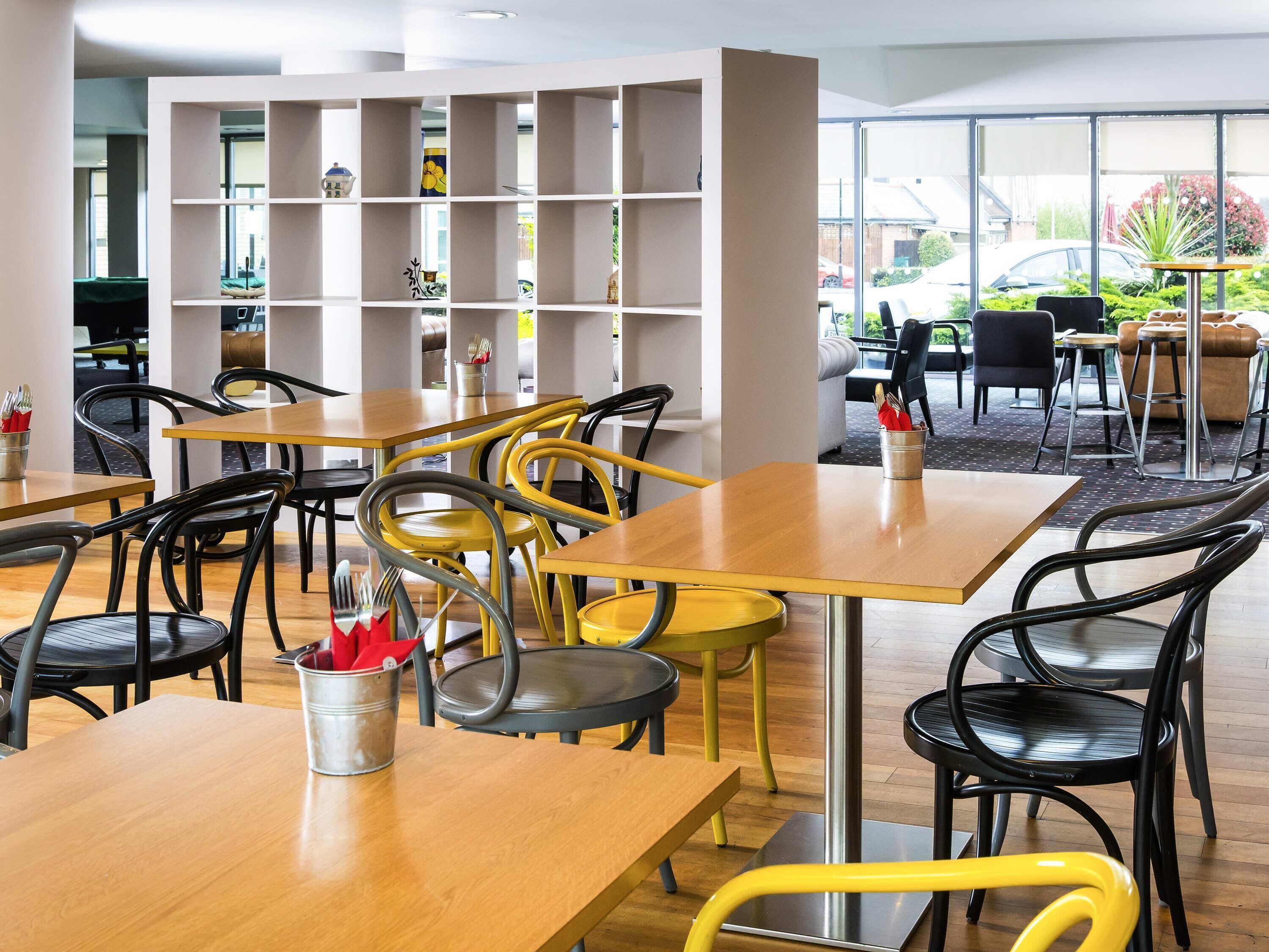 A modern dining area with wooden tables and chairs, a metal bookshelf, and a view of the outdoors through large windows.
