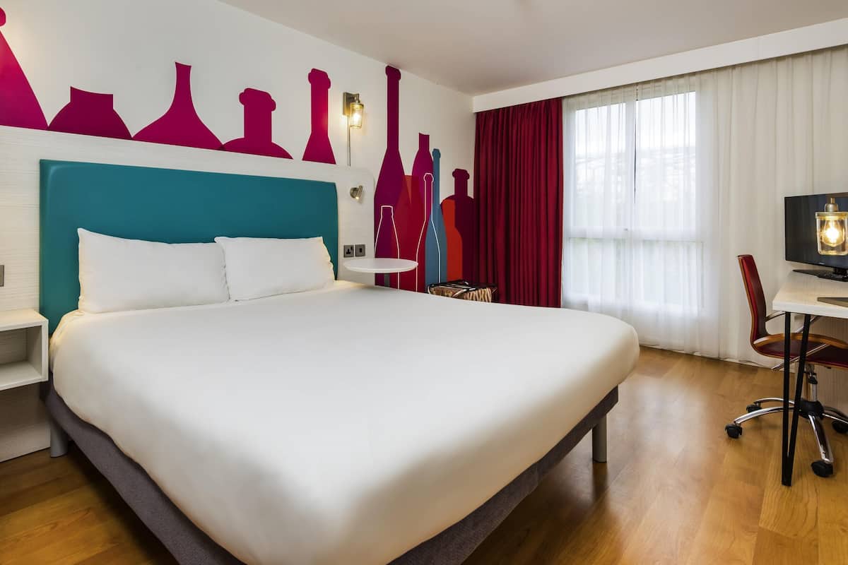 A modern hotel room with a large bed, a desk with a chair, a TV, and a wall with abstract art.