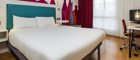 A modern hotel room with a large bed, a desk with a chair, a TV, and a wall with abstract art.