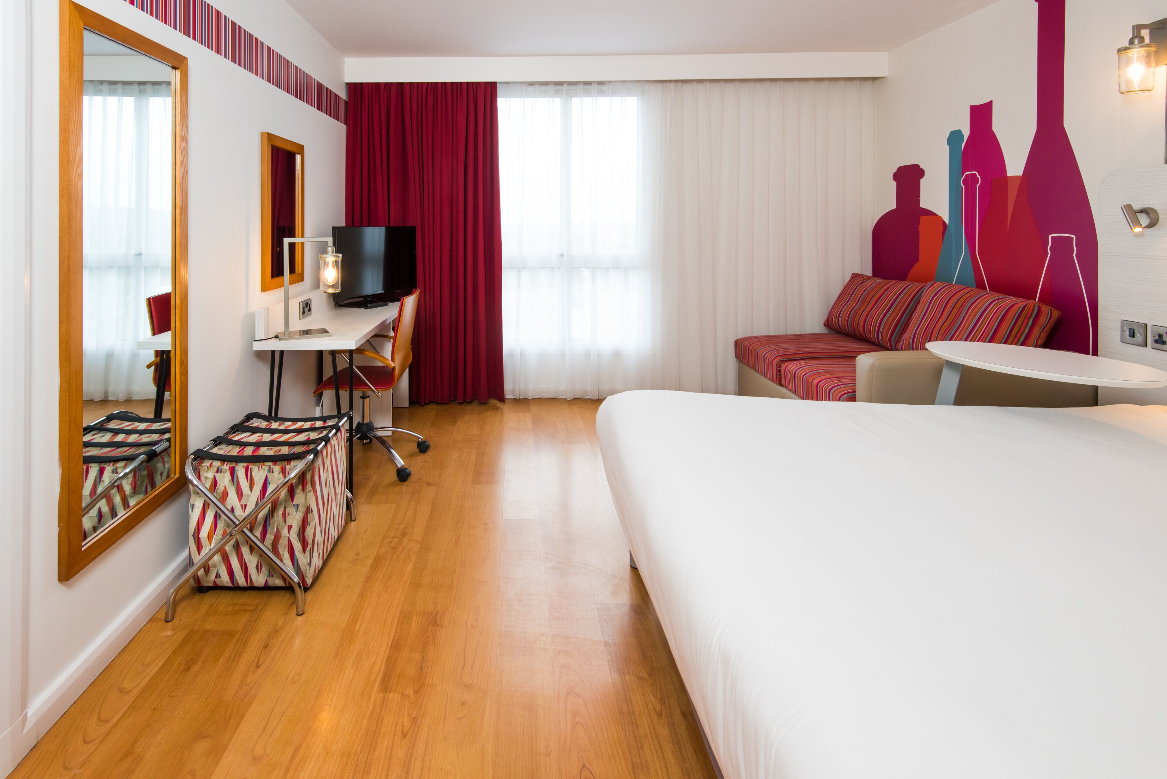 Ein Hotelzimmer mit Bett, Schreibtisch mit Computer, Stuhl, Koffer und Fernseher.