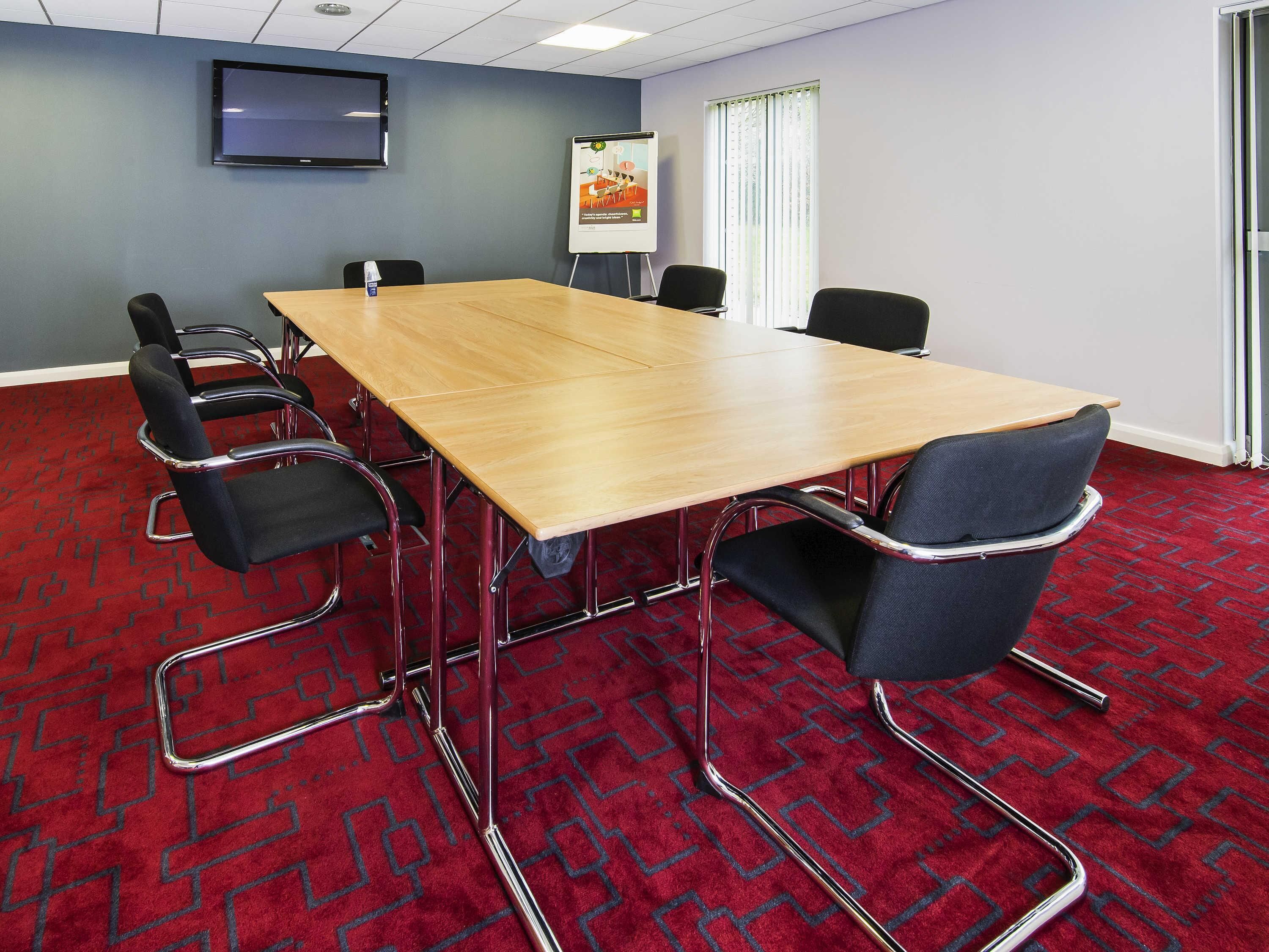 A conference room with a long table, six chairs, a projector screen, and a poster on the wall.