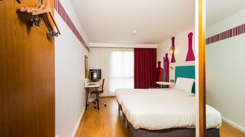 A hotel room with a wooden floor, a bed with white bedding, a desk with a chair, a television, and a window with red curtains.
