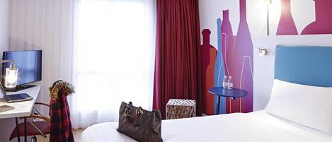 A hotel room with a bed, a desk with a laptop, a TV, a chair, a small table with bottles, and a decorative wall with silhouette cutouts.