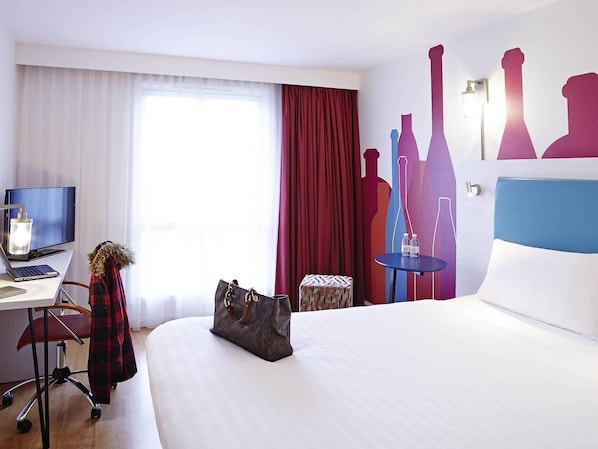 A hotel room with a bed, a desk with a laptop, a TV, a chair, a small table with bottles, and a decorative wall with silhouette cutouts. - Ibis Styles Barnsley Hotel (Barnsley)