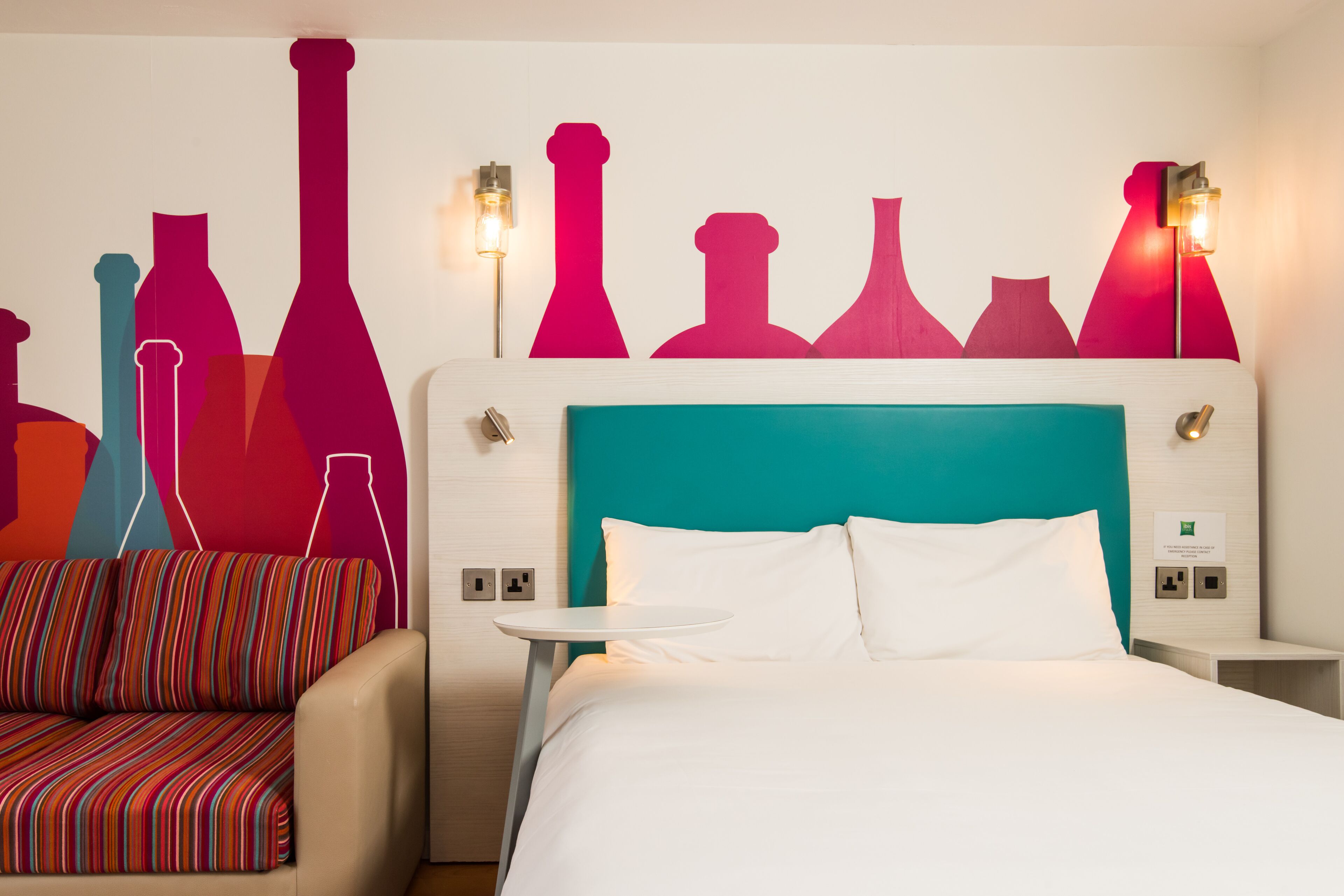 Une chambre d’hôtel avec une tête de lit de couleur turquoise, du linge de lit blanc, un canapé à rayures et une fresque murale représentant des formes abstraites.
