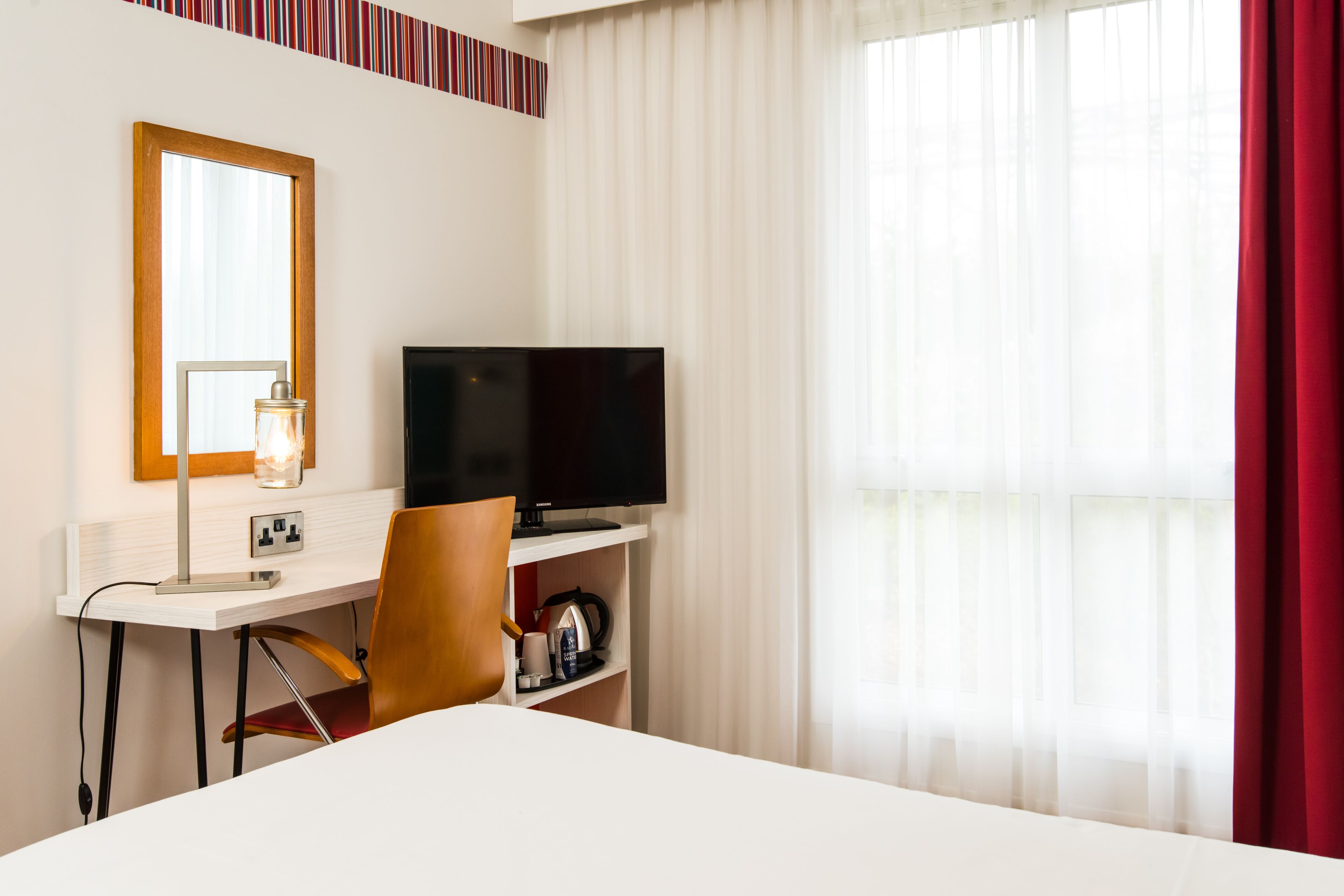 A hotel room with a bed, a desk with a television, a chair, a mirror, and a window with curtains.