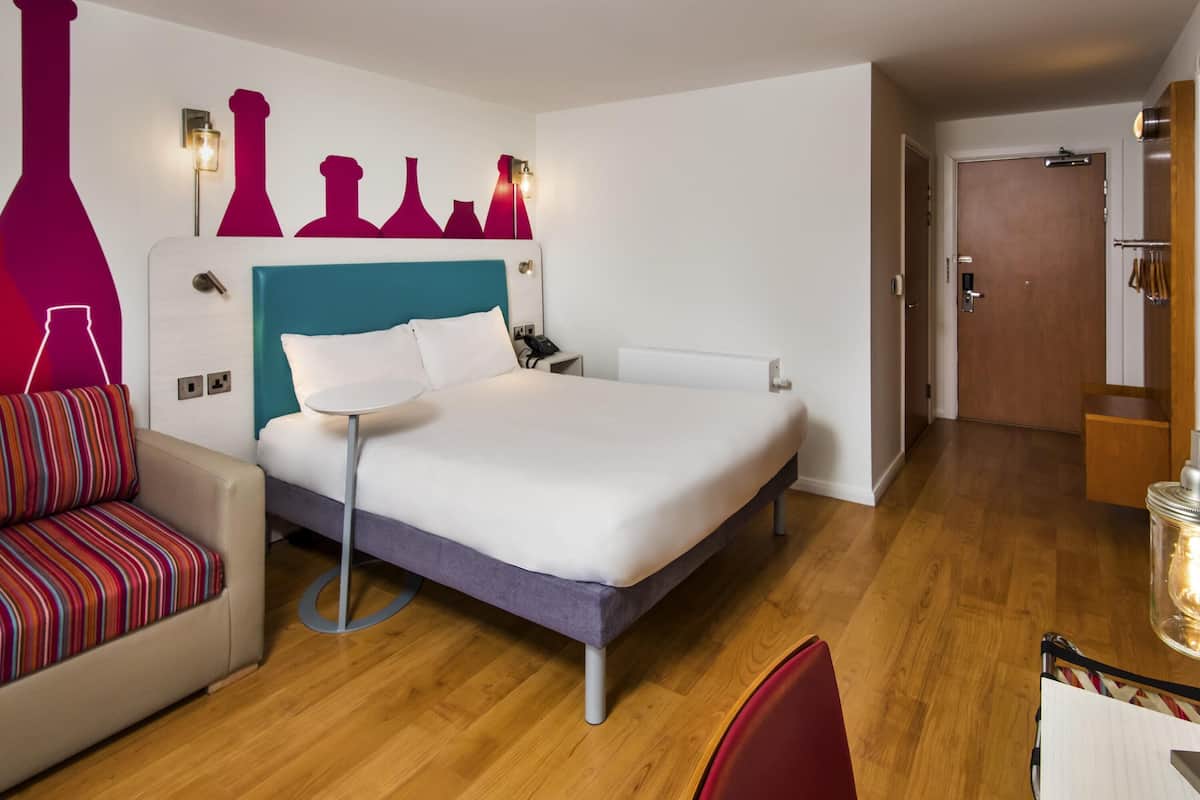 A hotel room with a bed, bedside table, chair, and a wall with decorative vases.