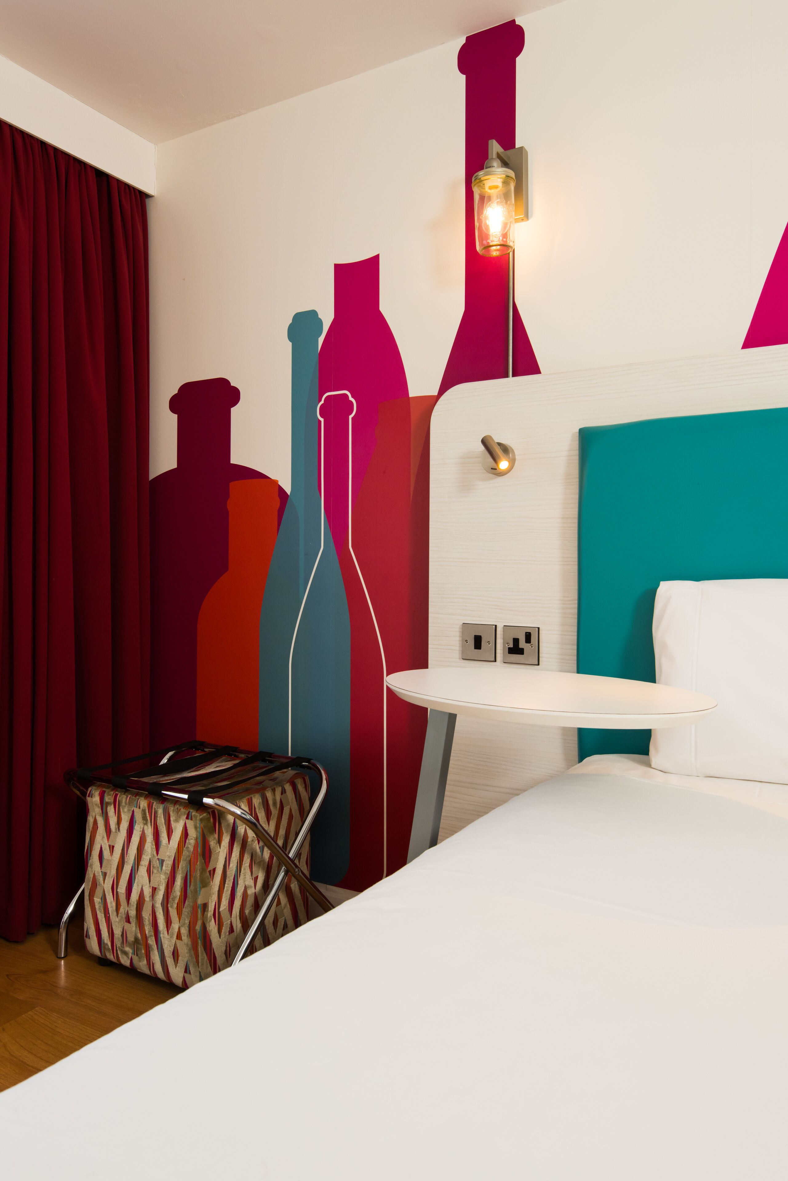 Habitación de hotel con cama, una mesita, pared con siluetas de botellas de colores, cortina roja y bolso estampado.