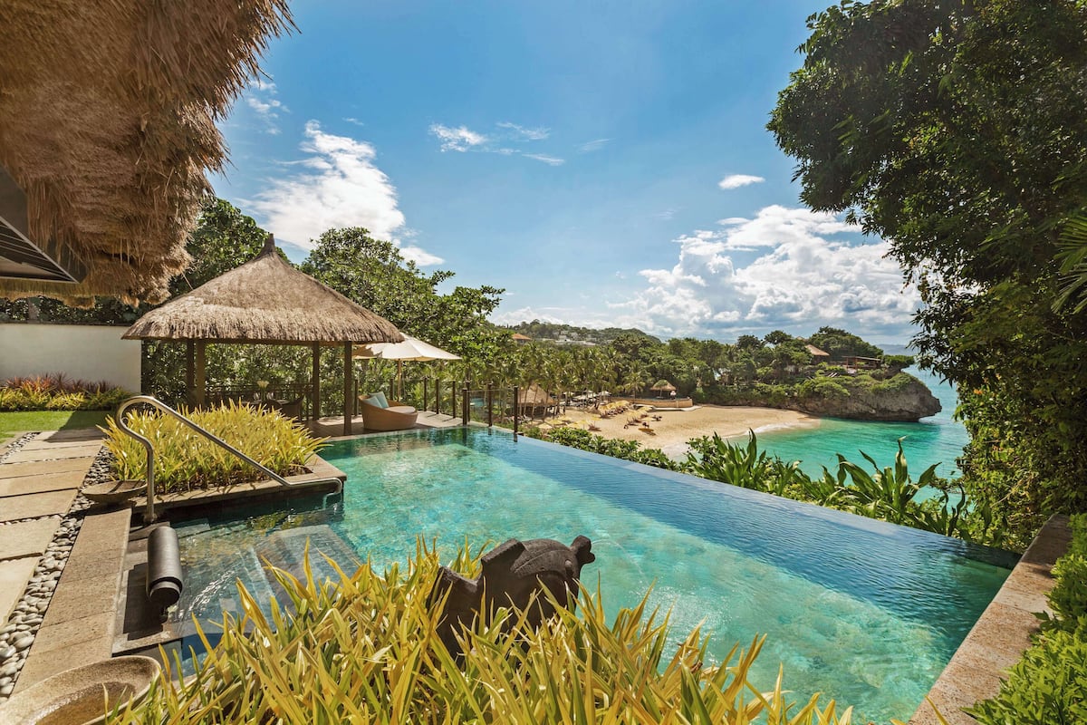 Ein Pool mit einem strohgedeckten Bauwerk, einem Steinweg und Blick auf einen Strand und Bäume.