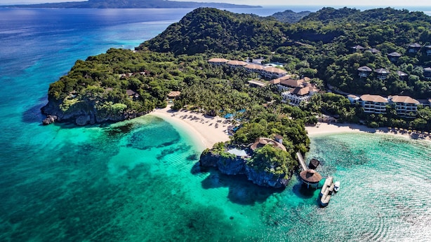 An aerial view of a tropical island with clear turquoise waters, a sandy beach, and a resort area with buildings and lush greenery.
