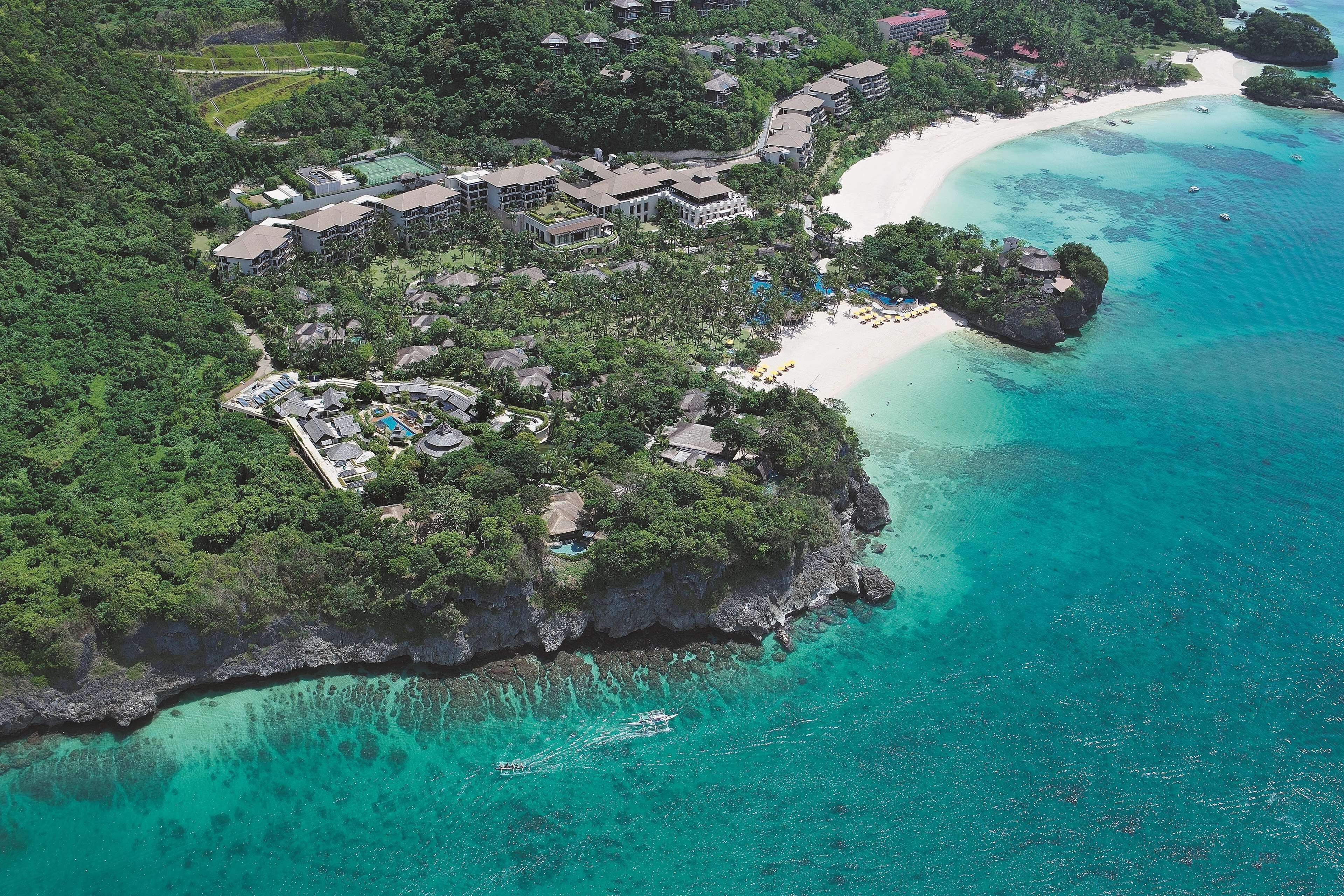 Luftaufnahme eines Resorts mit mehreren Gebäuden, einem Strand und klarem türkisblauem Wasser.