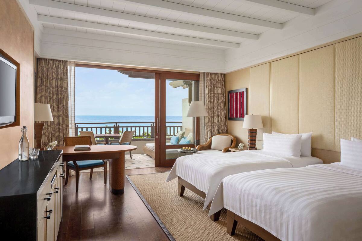 Ein Hotelzimmer mit zwei Betten, einem Essbereich, einem Fernseher und einem Balkon mit Meerblick.