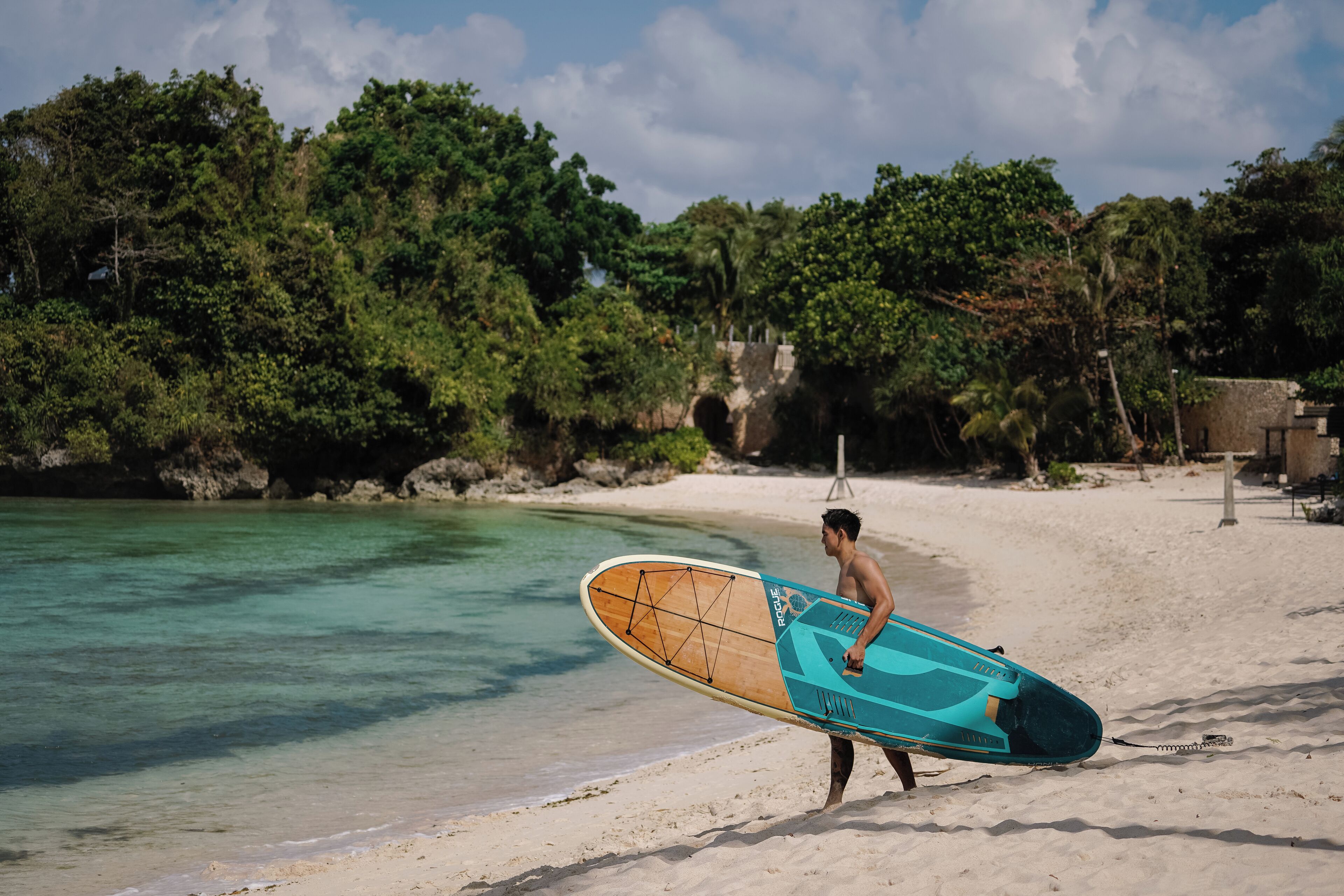 Una persona lleva una tabla de surf en una playa arenosa, con aguas cristalinas y abundante vegetación de fondo.