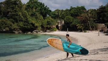Una persona lleva una tabla de surf en una playa arenosa, con aguas cristalinas y abundante vegetación de fondo.