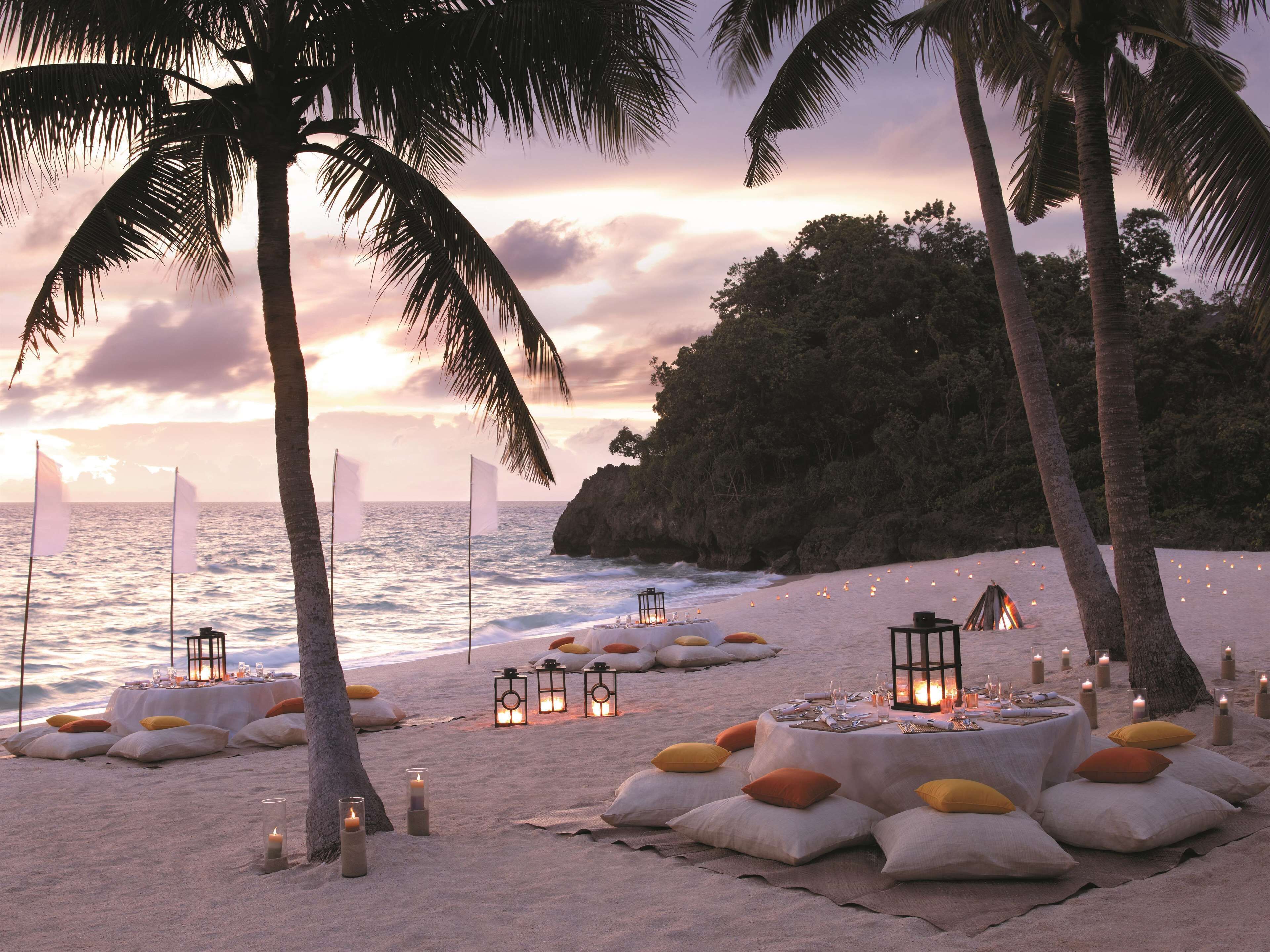 Un paisaje de playa con mesas dispuestas para cenar, rodeado de velas y palmeras.