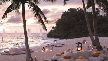 Un paisaje de playa con mesas dispuestas para cenar, rodeado de velas y palmeras.