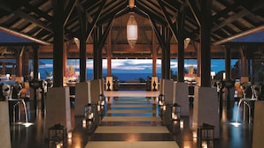 A spacious lobby with a view of the ocean, featuring a central staircase, wooden beams, and ambient lighting.