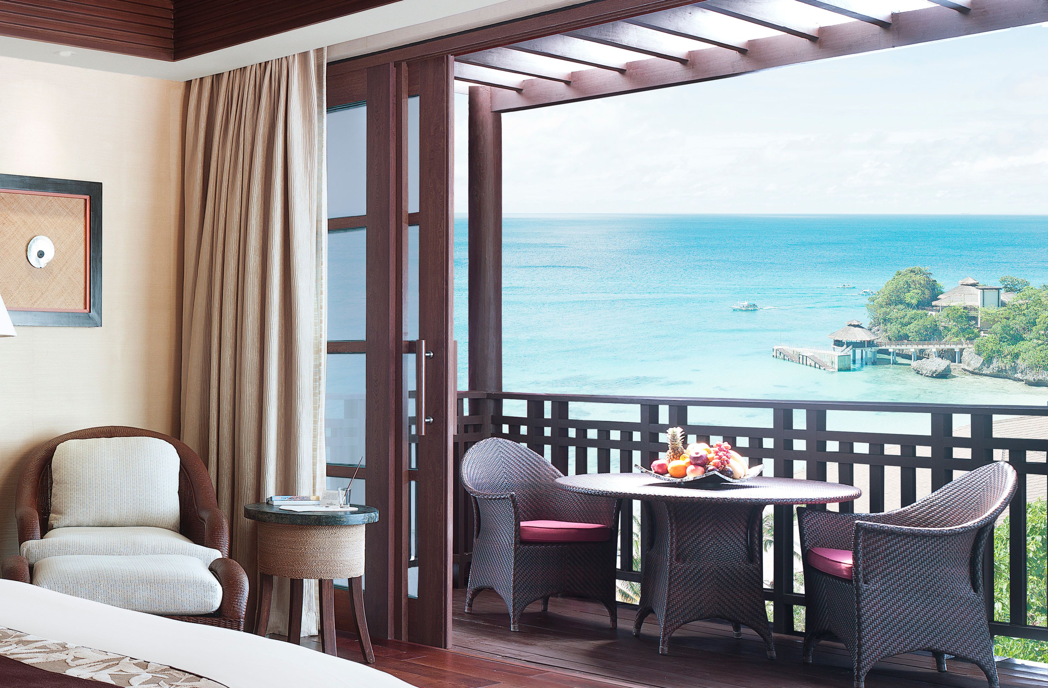 A balcony with wicker furniture, a table with fruit, and a view of the sea and distant buildings.