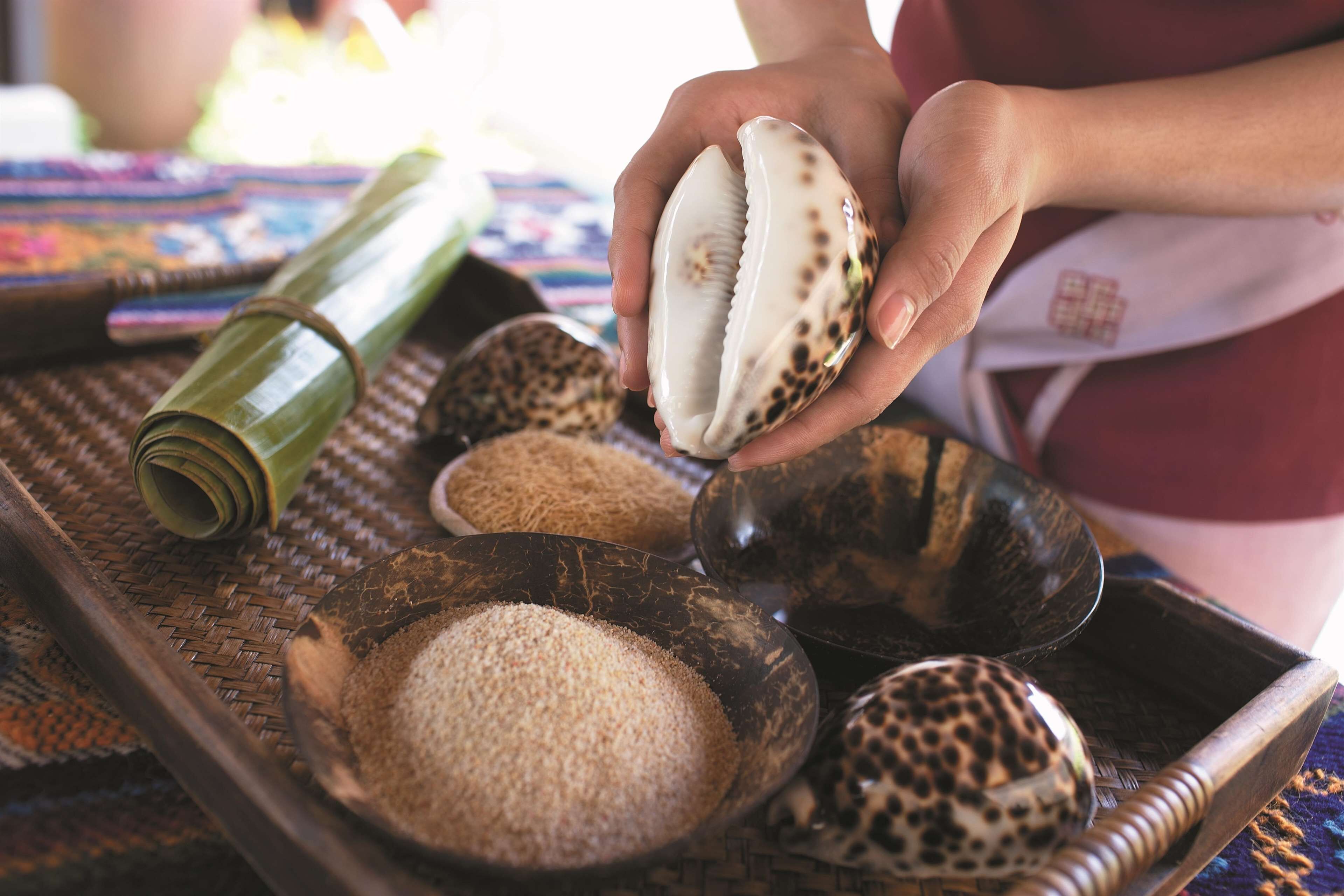 A person holding a shell, with bowls of grains and a woven mat on a tray.