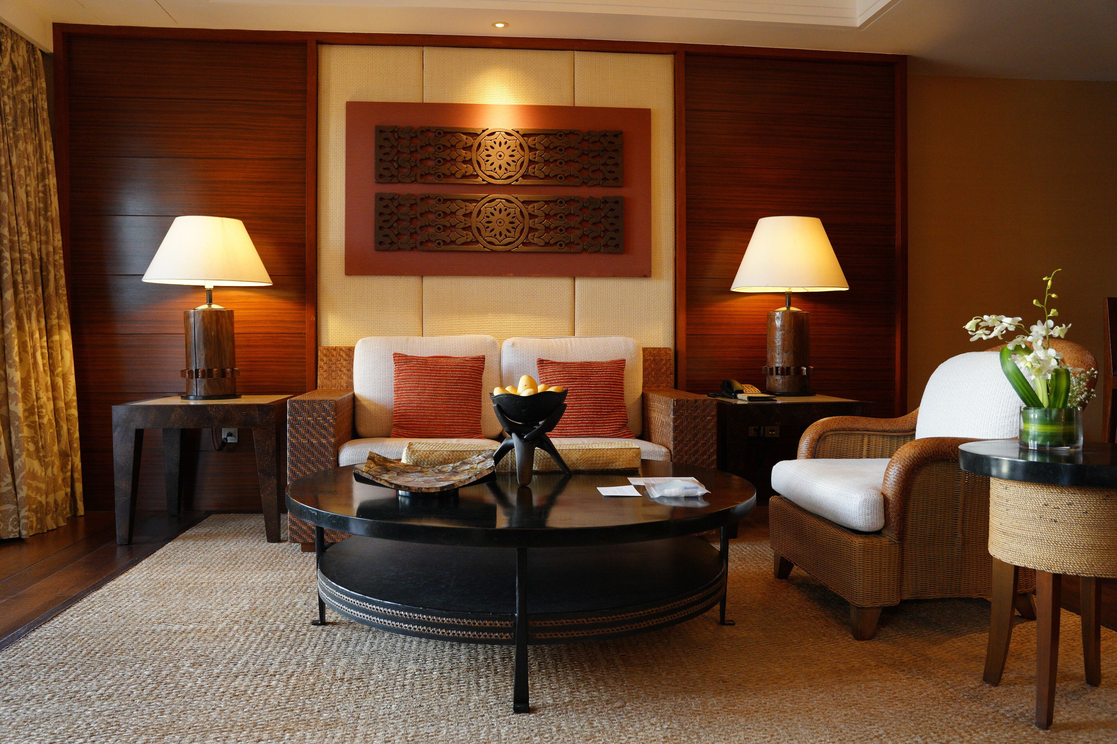 A hotel room with a round black coffee table, two wicker chairs, a sofa with red cushions, and two lamps on side tables.