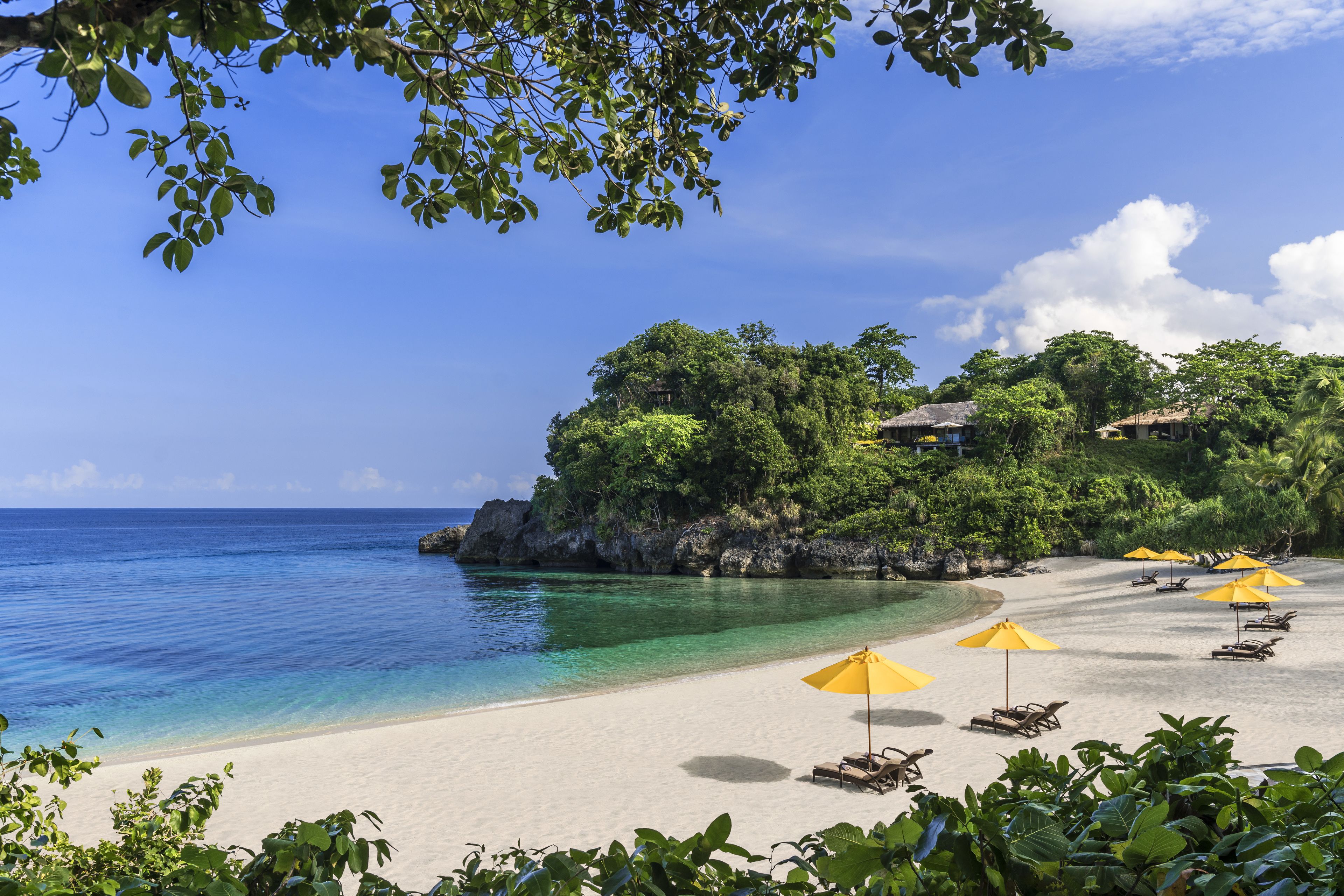 A beach with clear turquoise water, white sand, and yellow umbrellas. There are lounge chairs and a building on a small island in the background.