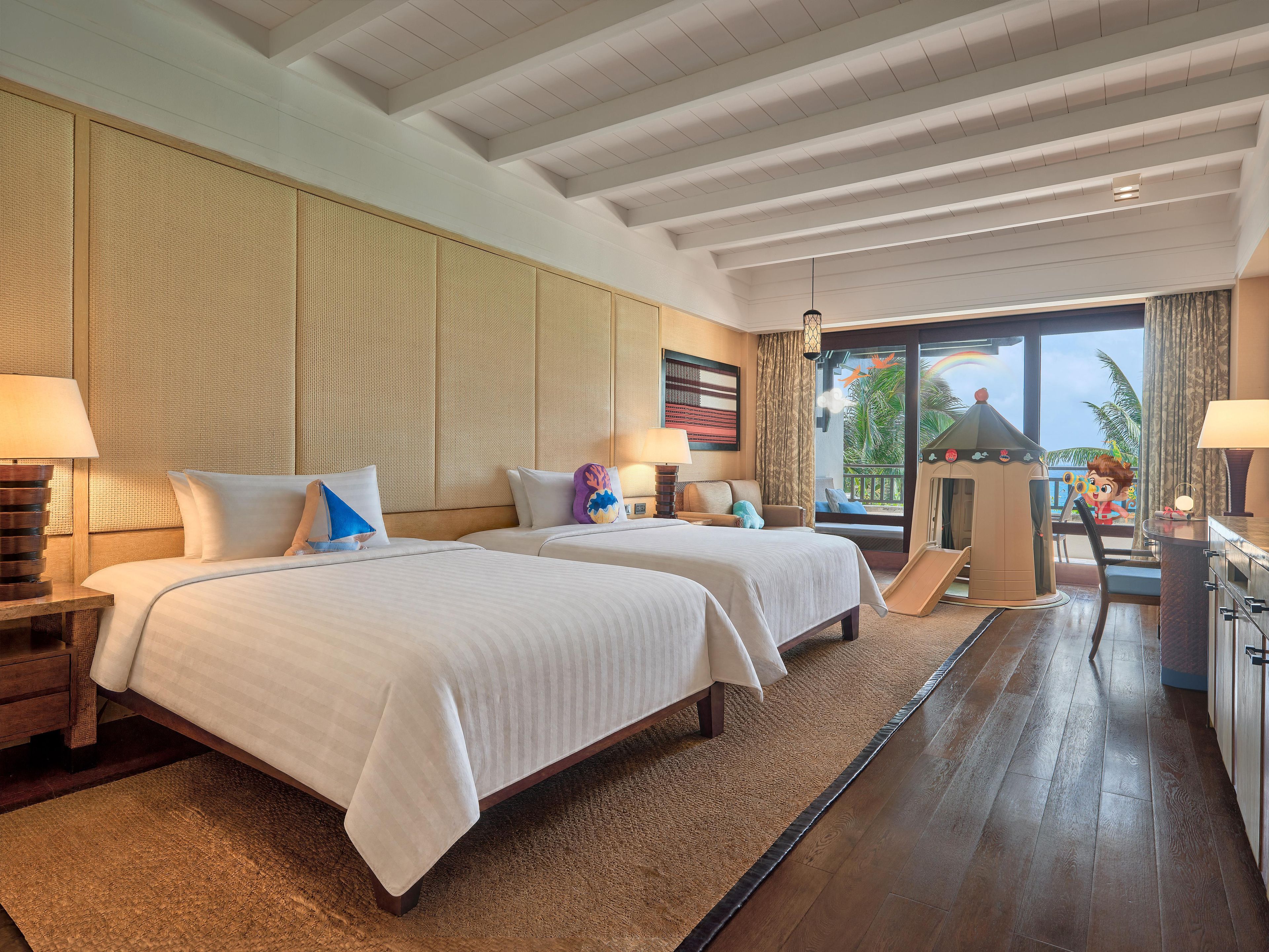 A hotel room with two beds, a desk, a chair, a play area, and a view of the outdoors.