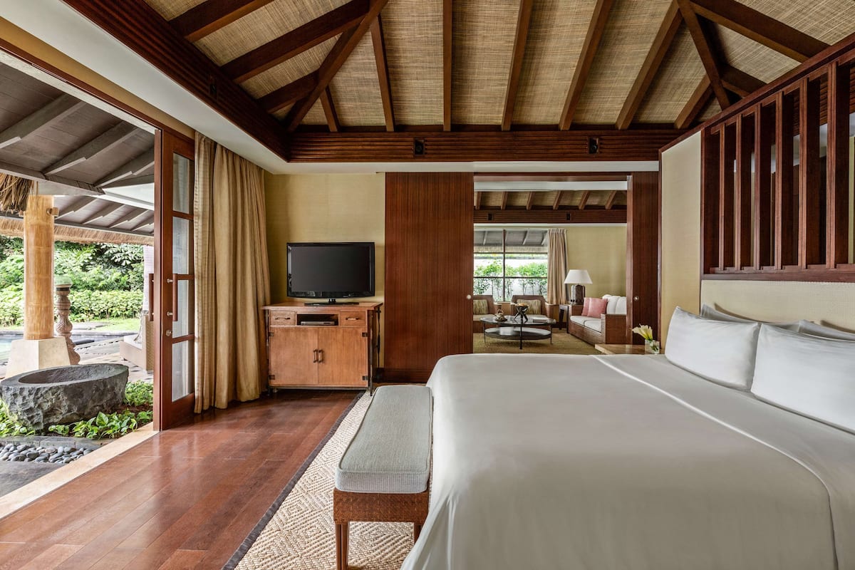 Ein geräumiges Schlafzimmer mit einem großen Bett, Holzbalkendecke, einem Fernseher auf einer Holzkommode und Blick auf einen Garten durch große Fenster.