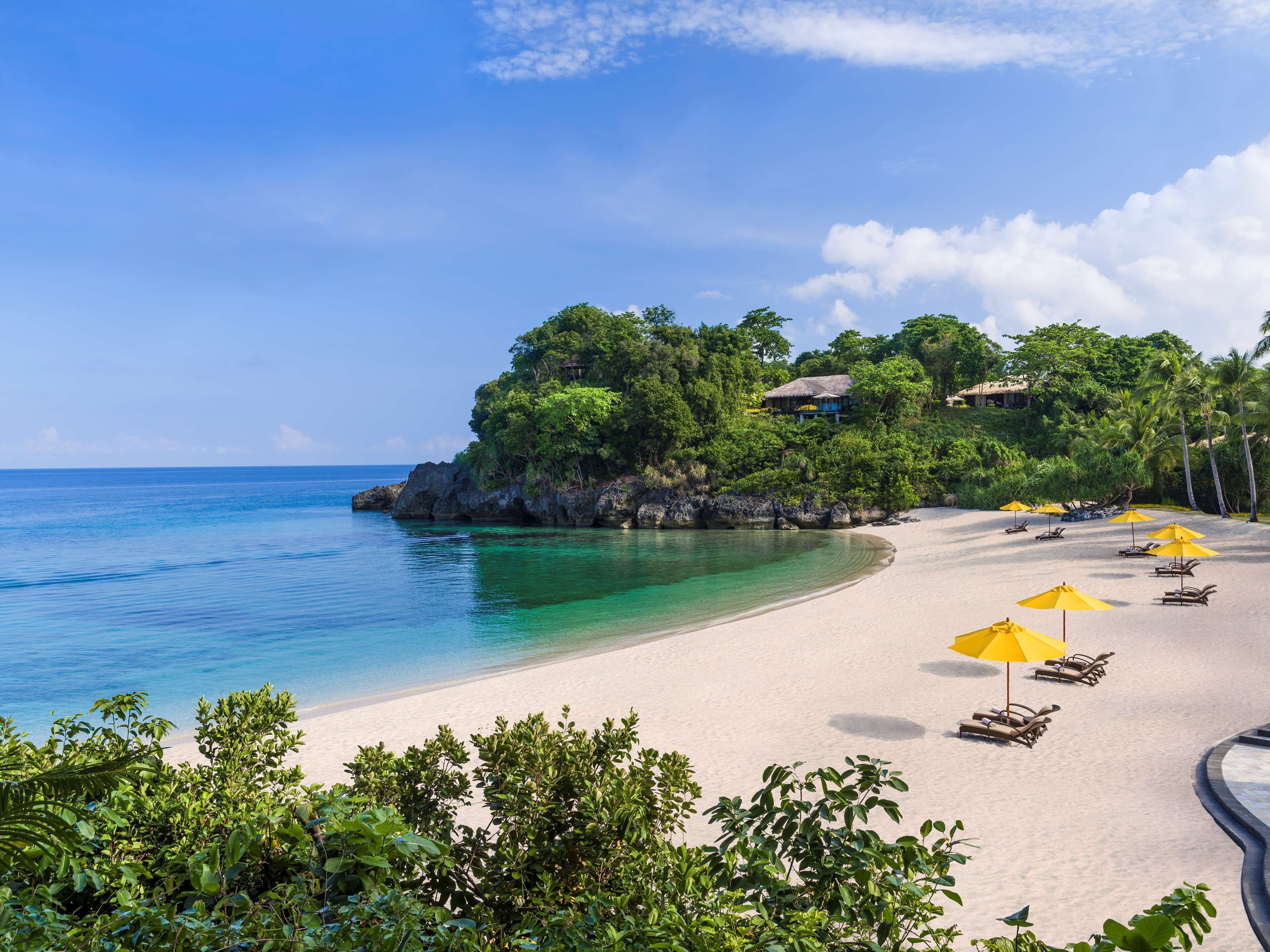 A beach with clear turquoise water, a sandy shore, and several yellow umbrellas. There are lounge chairs and a building with a thatched roof on a small island.