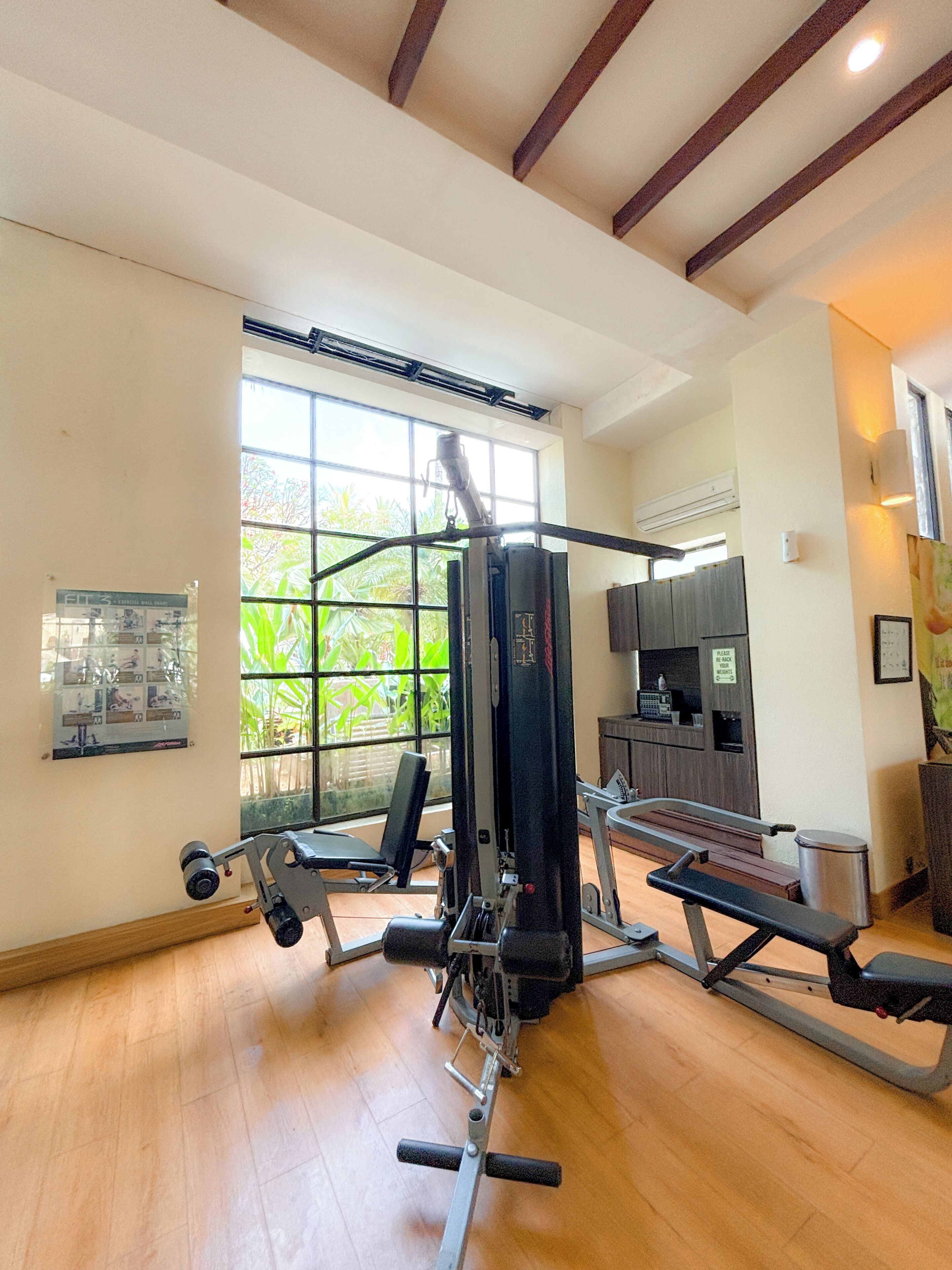 A gym with exercise equipment, a wooden floor, a large window with greenery outside, and a wall-mounted TV.
