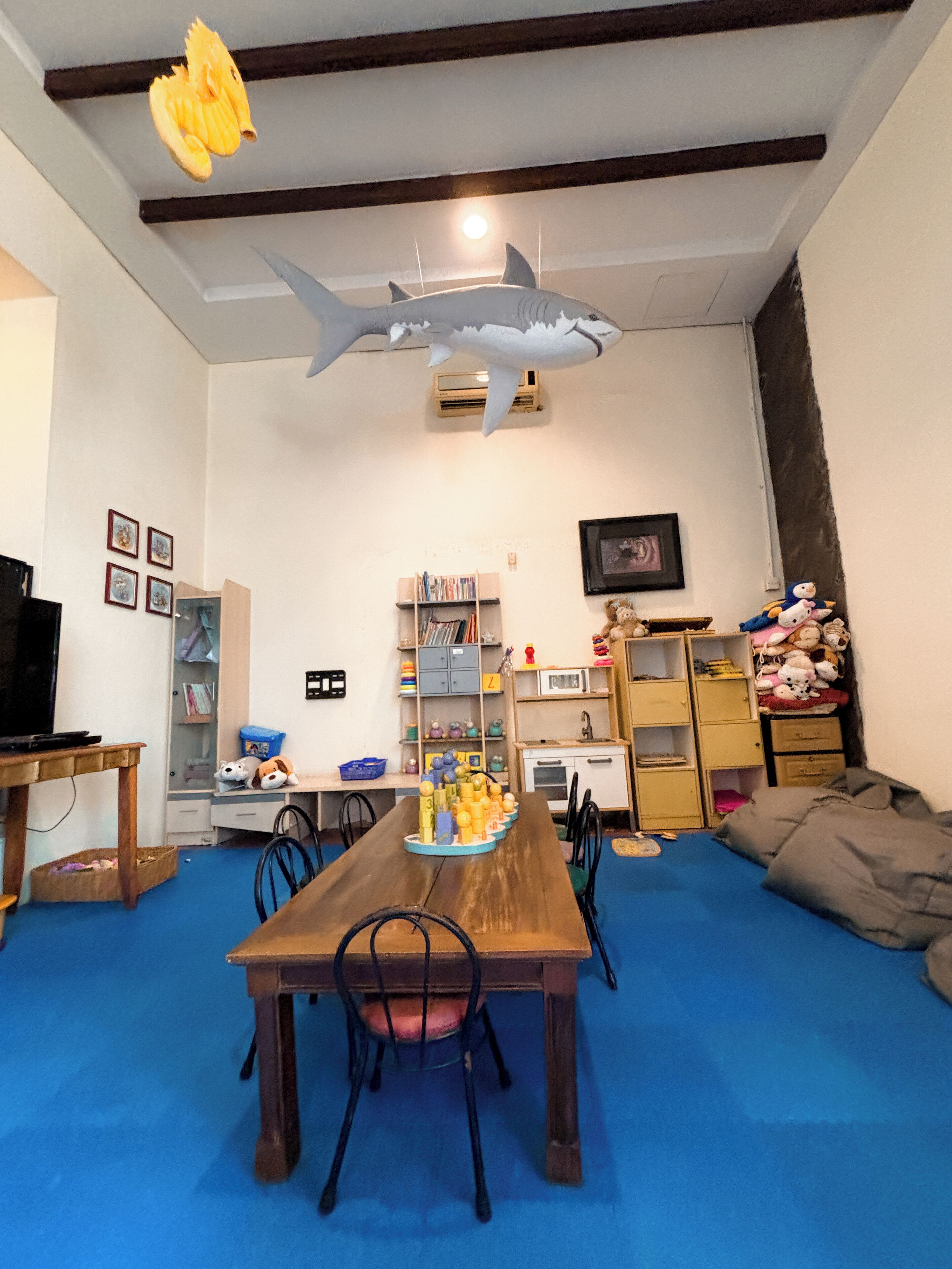 A room with a wooden dining table, chairs, a bookshelf, a television, and a shark decoration on the ceiling.