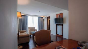 A hotel room with a dining table, two armchairs, a sofa, a TV, and a minibar.