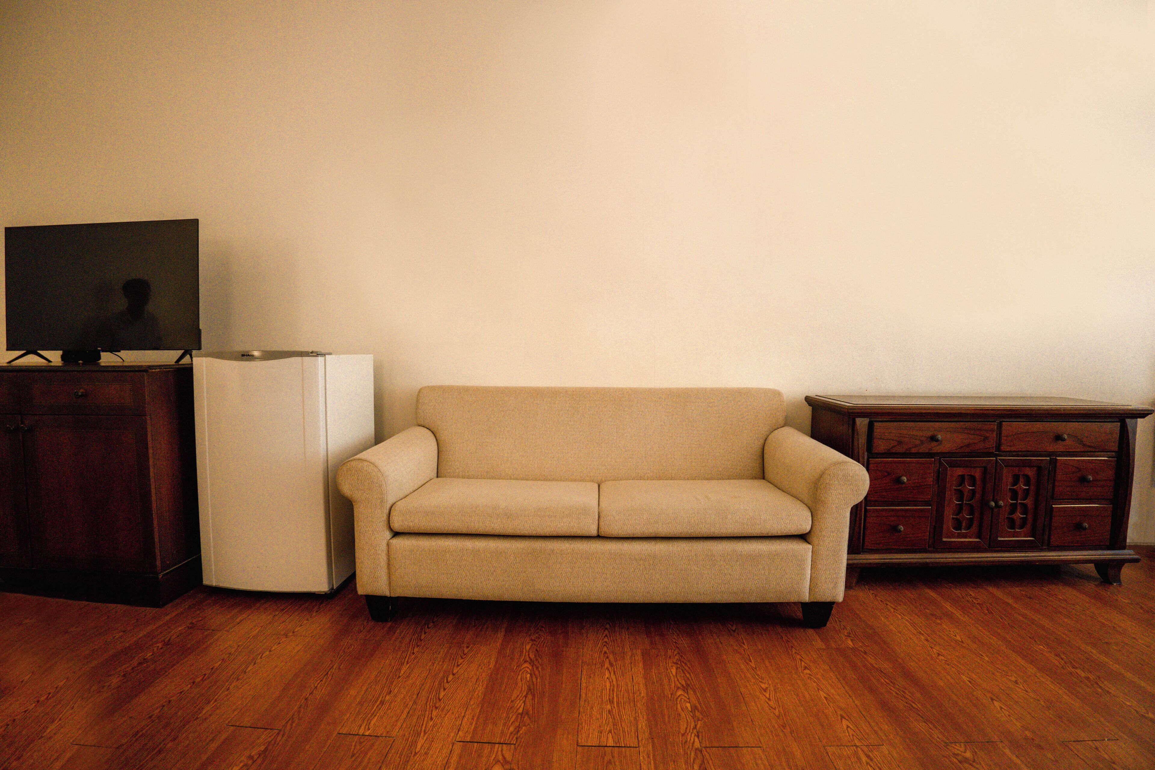 Une pièce avec un canapé beige, un réfrigérateur blanc, un meuble en bois et un téléviseur posé sur un meuble.