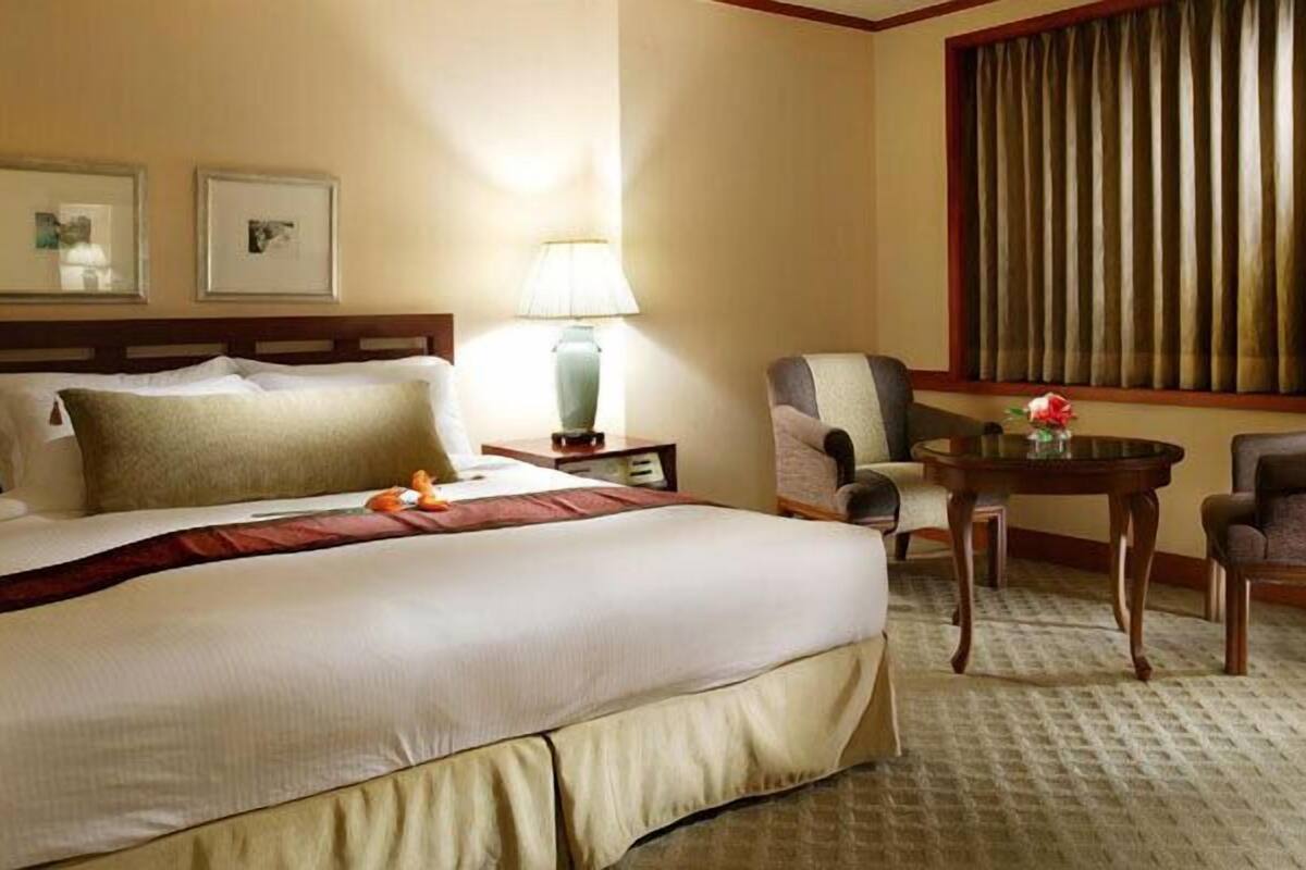 A hotel room with a large bed, a desk with a lamp, two chairs, and a window with curtains.