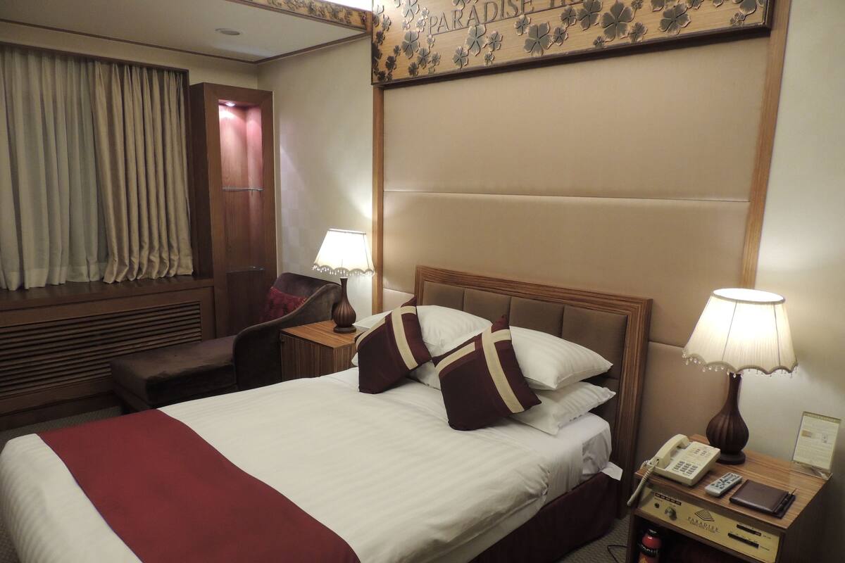 A hotel room with a large bed, a desk with a lamp, a telephone, and a small cabinet.