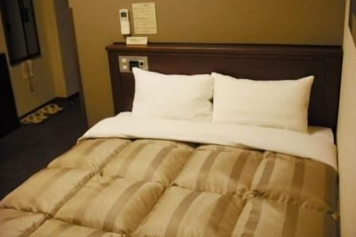 Hotellihuone, jossa on sänky, puinen pääty, seinään kiinnitetty puhelin ja pieni pöytä, jonka päällä on ruokalista tai tiedote.