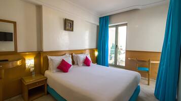 Habitación de hotel con una cama grande, una mesita de noche de madera, una pequeña mesa, un espejo, un cuadro en la pared y vistas al exterior a través de una ventana con cortinas azules.